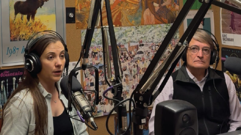 A woman and a man wear headphones. The woman sits in front of a microphone.