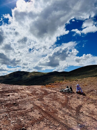 A cyclist sits by her bike on a sunny day on top of a mountain.