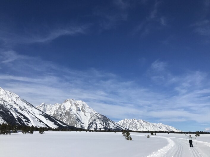 A cross-country skier stands with their arms extended on a groomed road that’s covered in snow. In the background are tall mountain peaks also covered in snow.