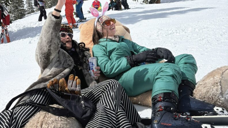 Two women in crazy outfits lounge on a massive teddy bear on a ski slope.