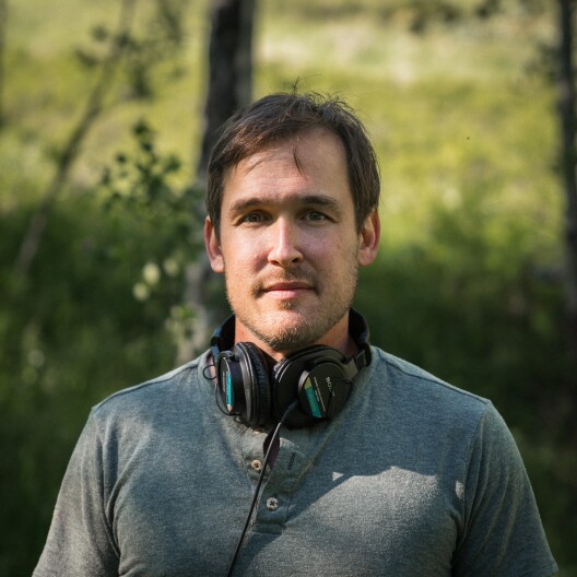 A man with brown hair wears headphones around his neck. There’s a blurred forest background.