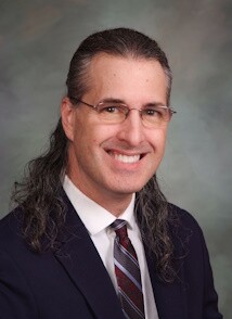 A headshot of a man with long hair and glasses in a suit.