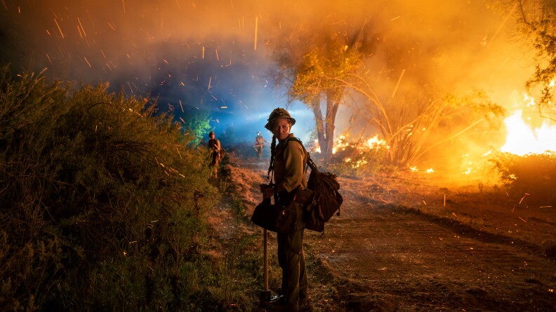 A woman in wildland firefighting gear turns and looks directly at the camera as a fire rages beside her.