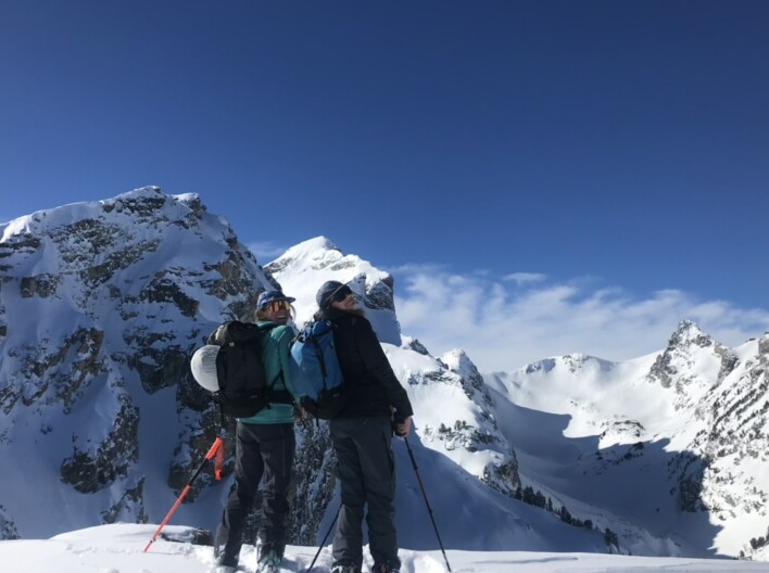 Two women with skis and big backpacks stand on top of a peak and smile at the camera over their shoulders. Behind them are snow-covered mountains and a blue sky.