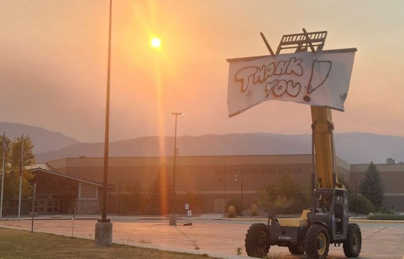 A crane holds up a sheet that's been spray painted with the message, "Thank you!" The crane is in the parking lot for Tongue River High School. A smokey sunset washes the scene in an eerie red glow.