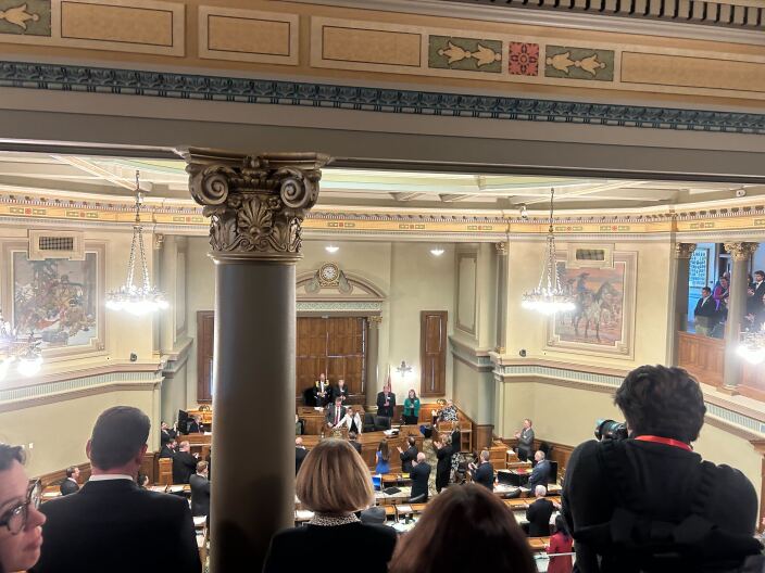 Looking over press on a balcony into an ornate chamber where members of the Wyoming Legislature stand.