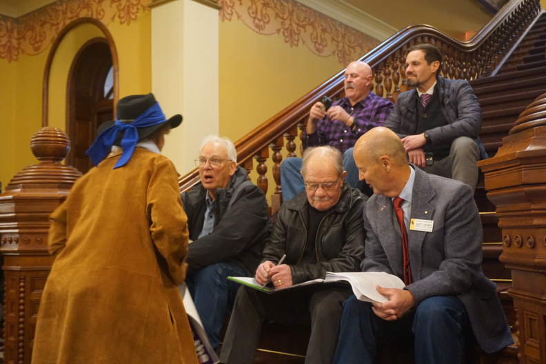 A group of men sit on stairs talking as one stands in front of them.