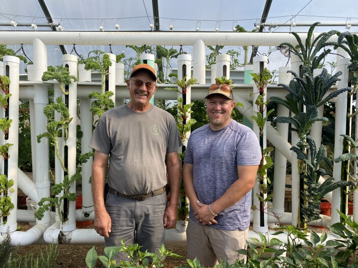 An older and younger man wearing baseball hats and t-shirts pose in front of a hydroponic greenhouse where kale and Swiss chard grow from a lattice-work of white PVC pipes.
