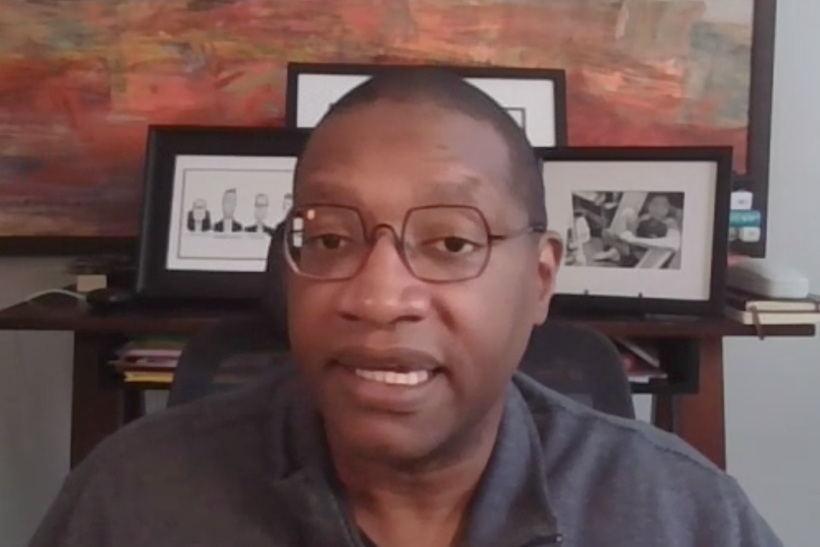 A screenshot from a Zoom call shows Kareem Crayton sitting in front of a stand with photos in frames and a painting hung on a wall. Crayton is a Black man with short black hair. He is wearing glasses and a gray shirt.