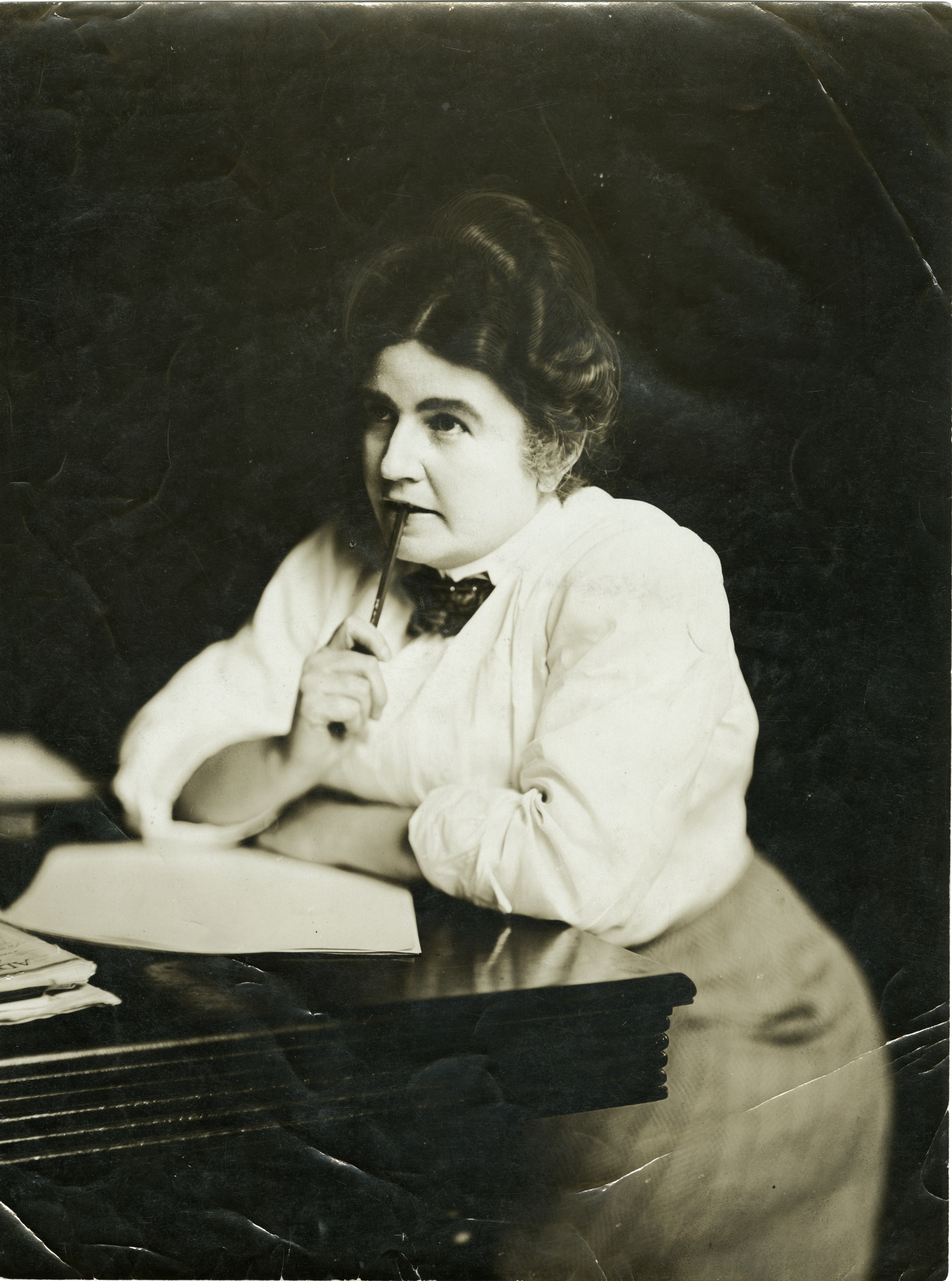 Black and white portrait of author Gene Stratton-Porter leaning on a wooden desk, holding a pen to her lips in a thoughtful pose.