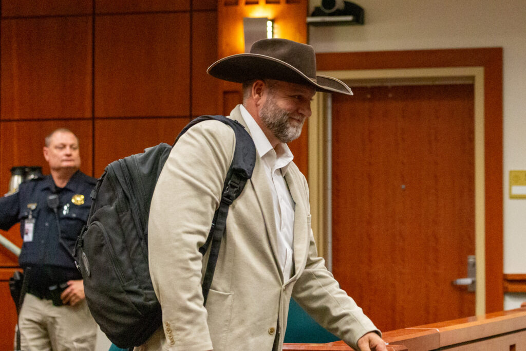 A man is seen walking wearing a brown cowboy hat, tan suit jacket, white shirt and a backpack slung over one shoulder. There is a police officer standing still in the background. 