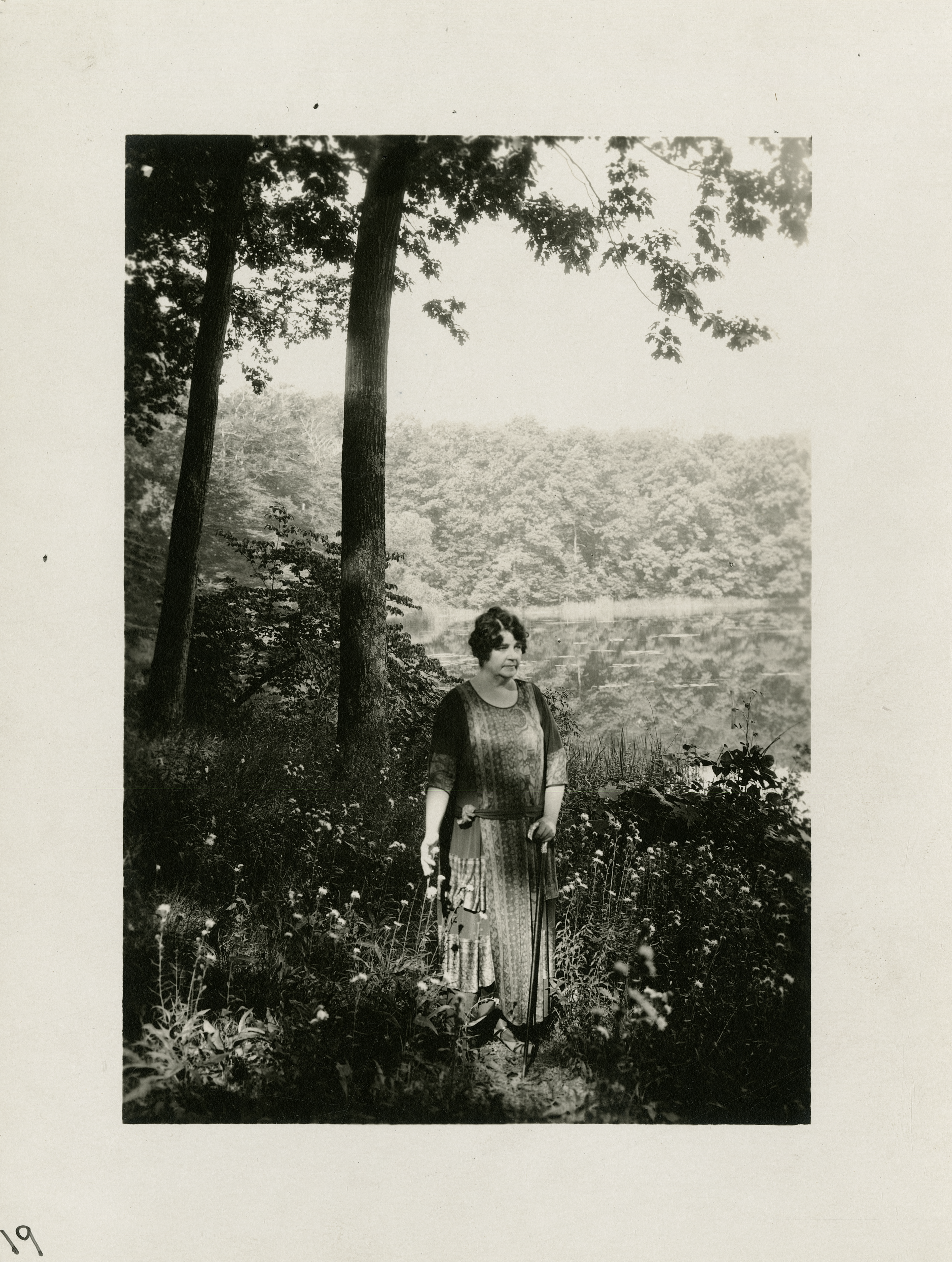 Black and white portrait of Gene Stratton-Porter standing in a wooded area near a lake, surrounded by wildflowers and trees.