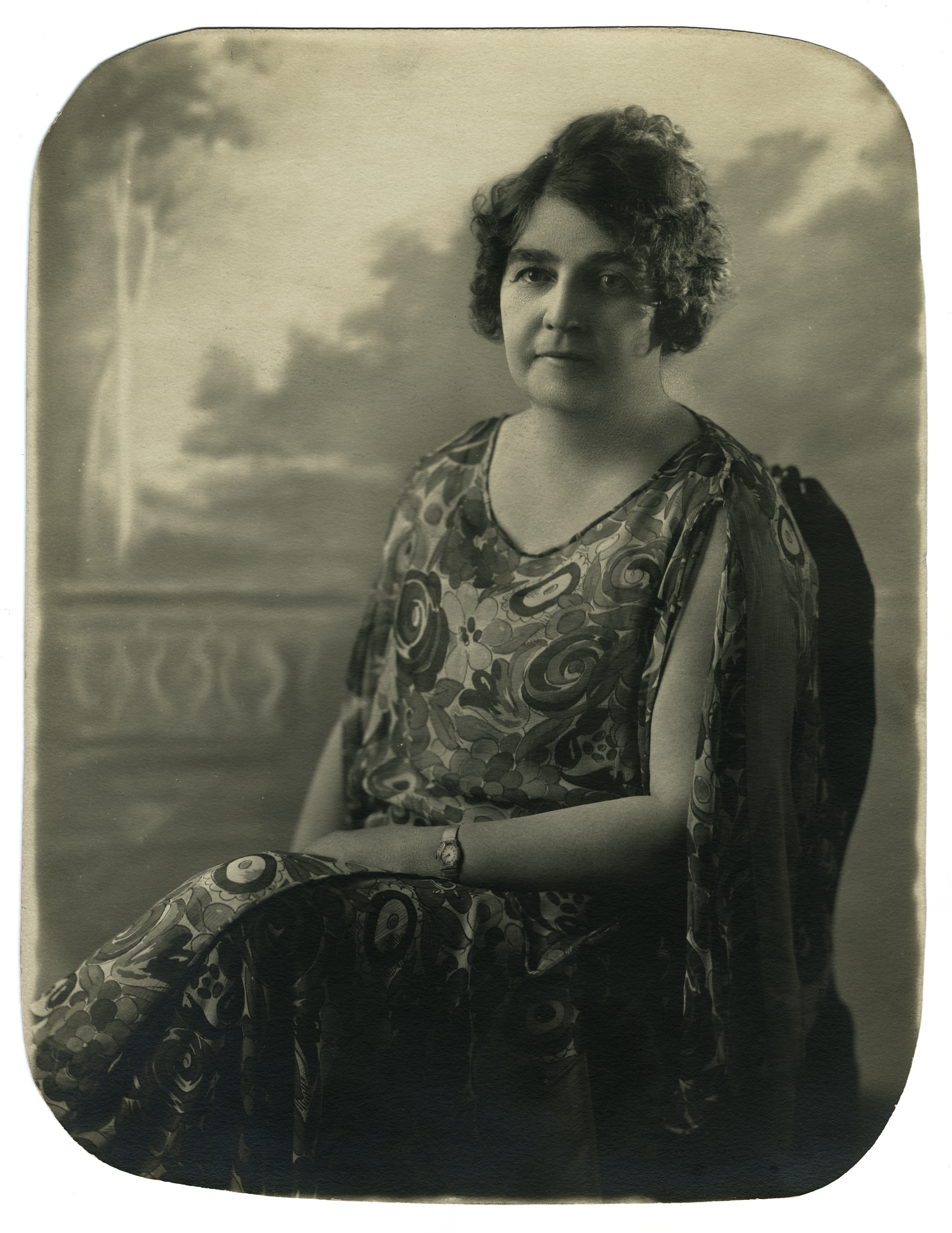 Sepia-toned studio portrait of author Gene Stratton-Porter with short, curled dark hair, wearing a patterned sleeveless tunic dress and seated against a painted backdrop of a cloudy sky and terrace.