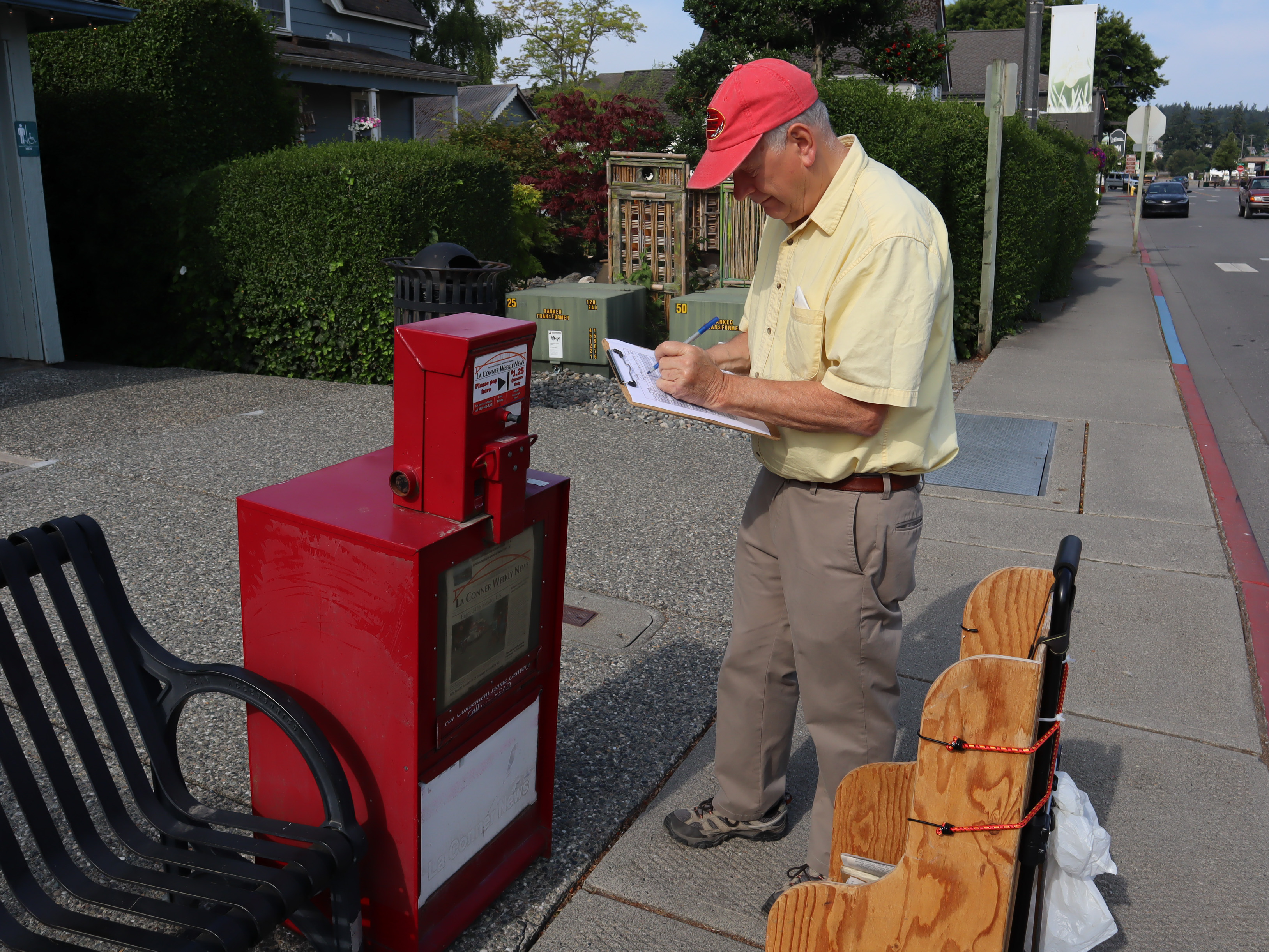 In a yellow shirt and tan hat, La Conner Weekly News' editor and publisher Ken Stern writes on a clipboard how many copies he has sold of last week's paper out of the red newspaper box he is restocking.