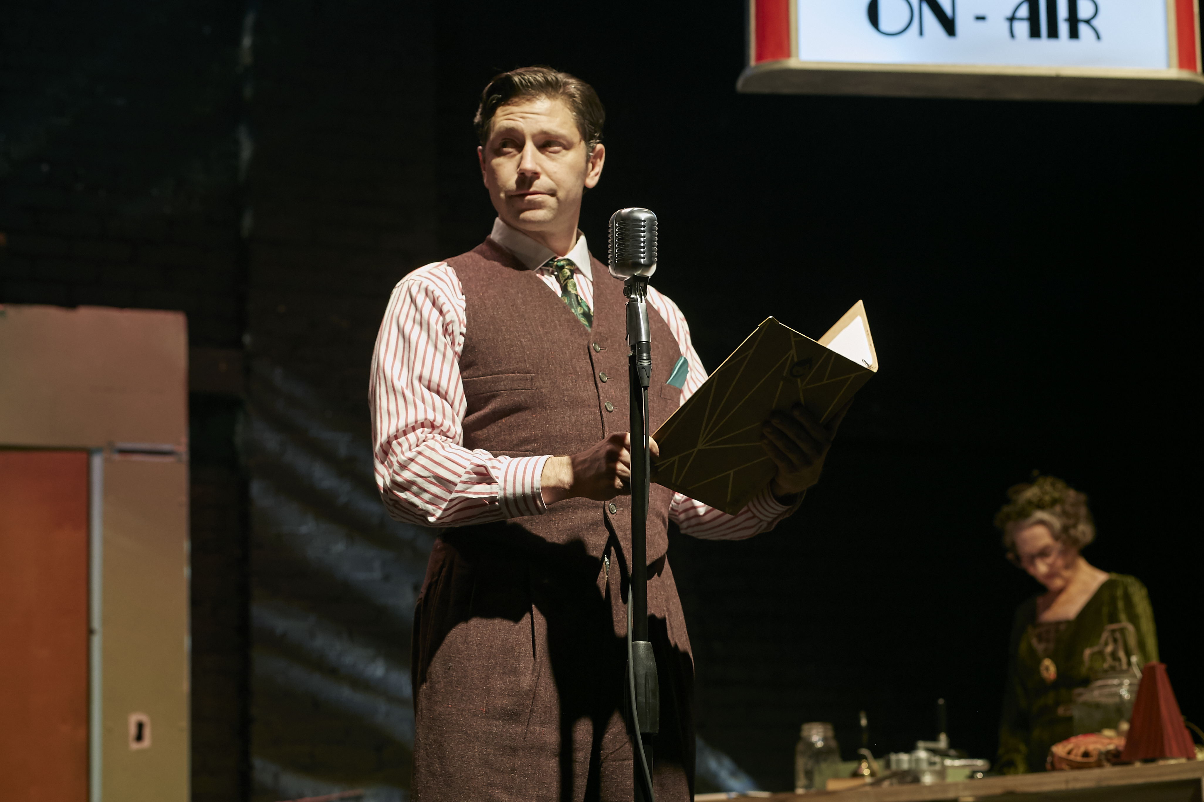 A performer in period clothing holds a radio script beside a vintage microphone