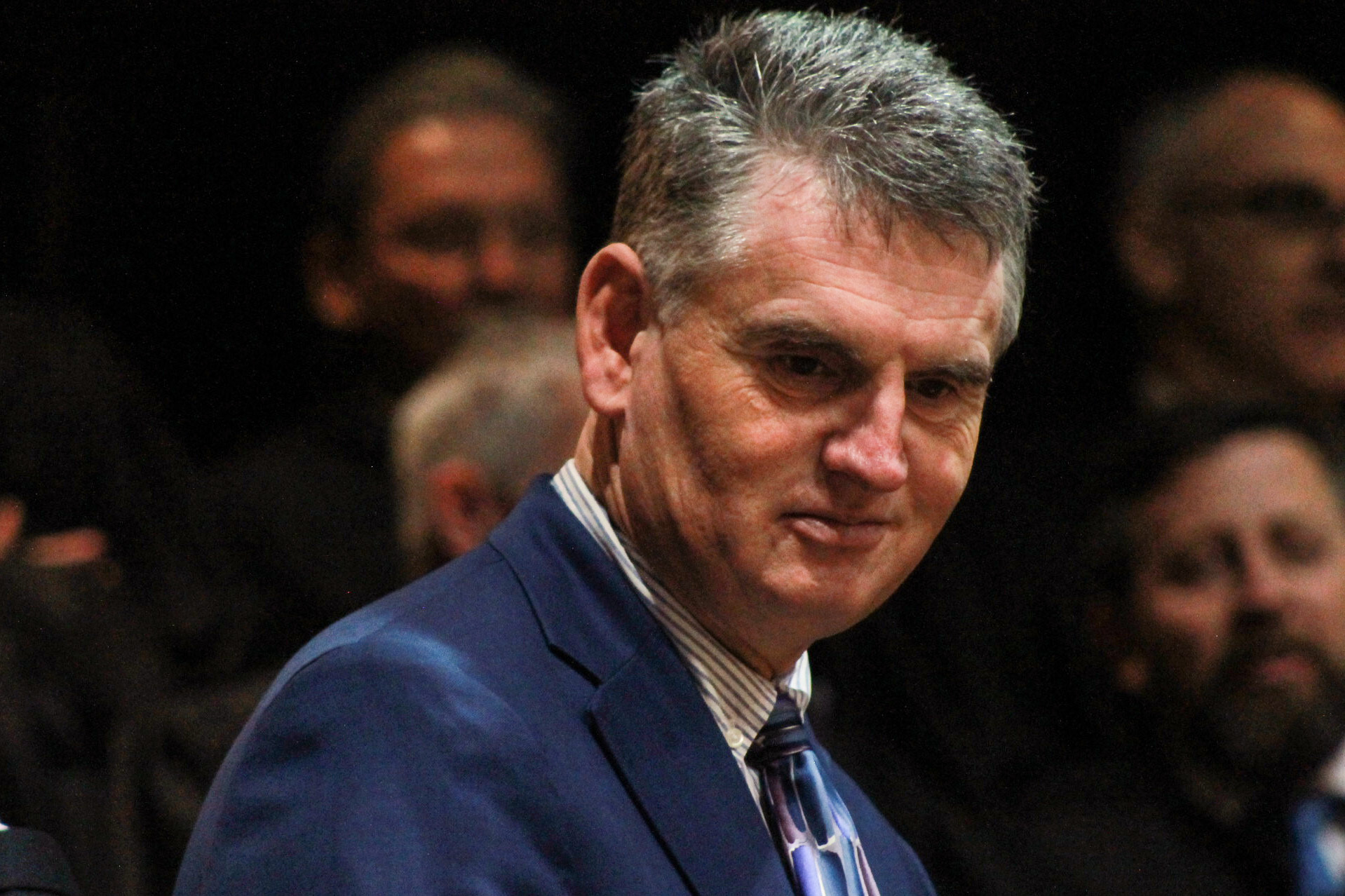 Daniel Elliott stands in the House chamber balcony, looking down. Elliott is a White man with gray hair. He is wearing a dark blue suit, a white shirt and a blue patterned tie. Behind him, out of focus, are people sitting in seats in the balcony.