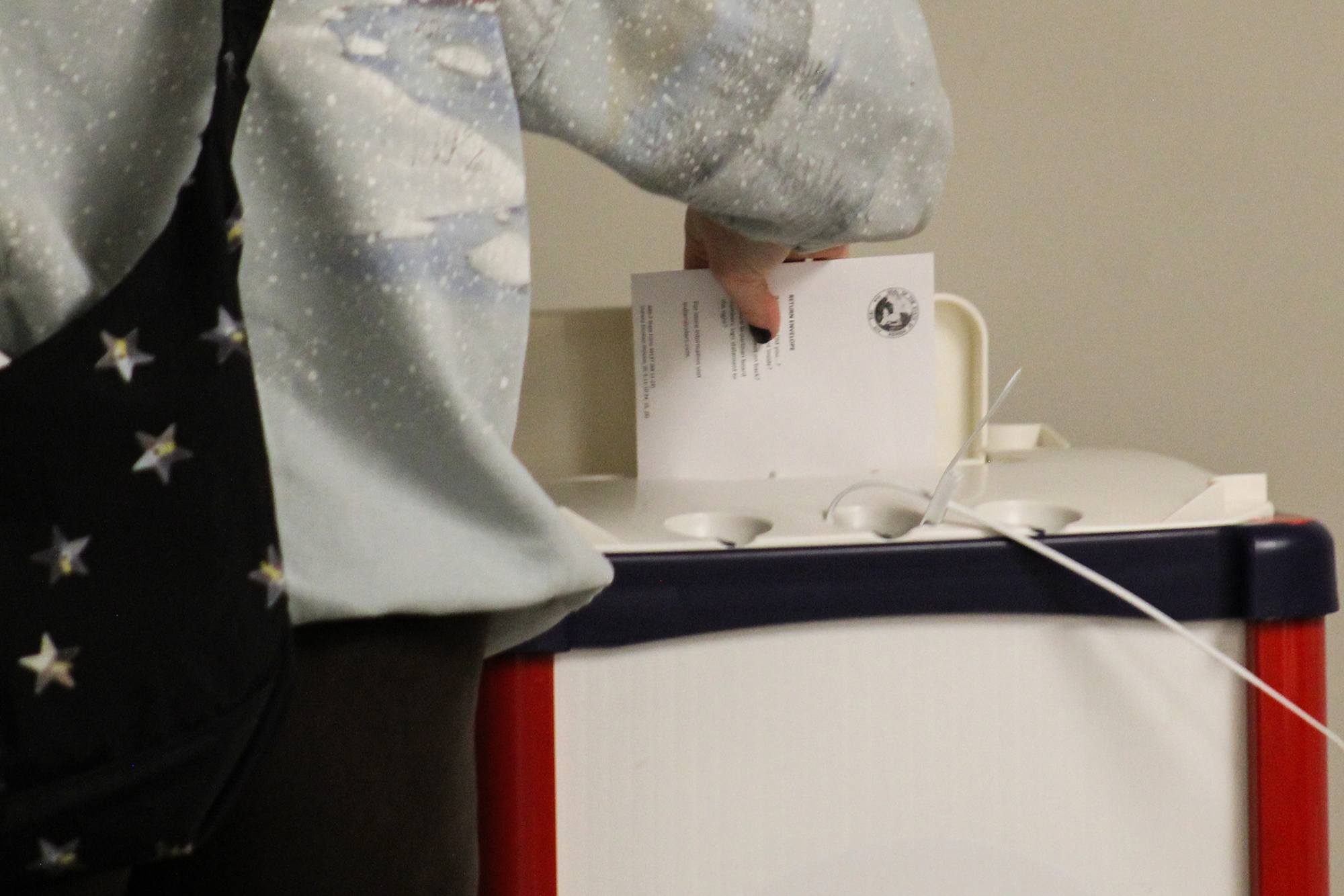 A person returns their ballot into a box at a vote center in Marion County. They wear a denim jacket and have a bag slung across their back with stars affixed to it.