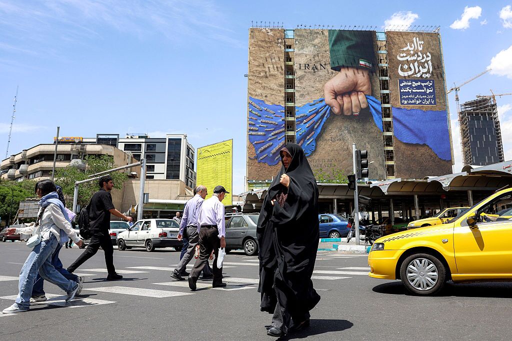 A woman crosses a street near a billboard on a building depicting the Strait of Hormuz with a caption in Persian reading "Forever in Iran