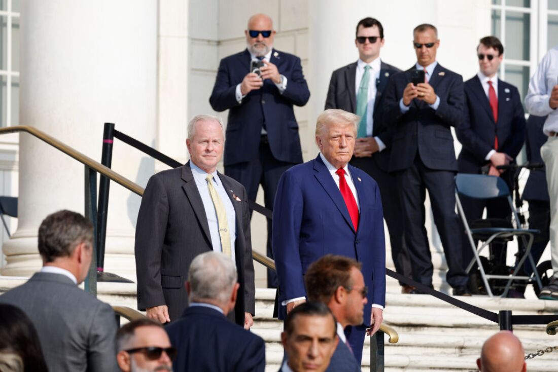 This photo shows former President Donald Trump at Arlington National Cemetery on Monday. He's wearing a blue suit and red tie and is standing outdoors on stairs while surrounded by various officials and staffers in suits.