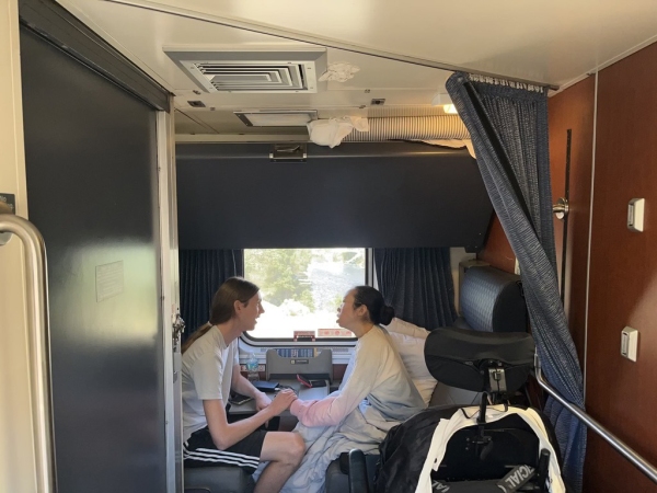 Peter and Aubrie in their accessible room on the California Zephyr Amtrak train. They sit next to a window facing each other. Aubrie’s wheelchair is to her side. Outside the window, water and trees rush by.