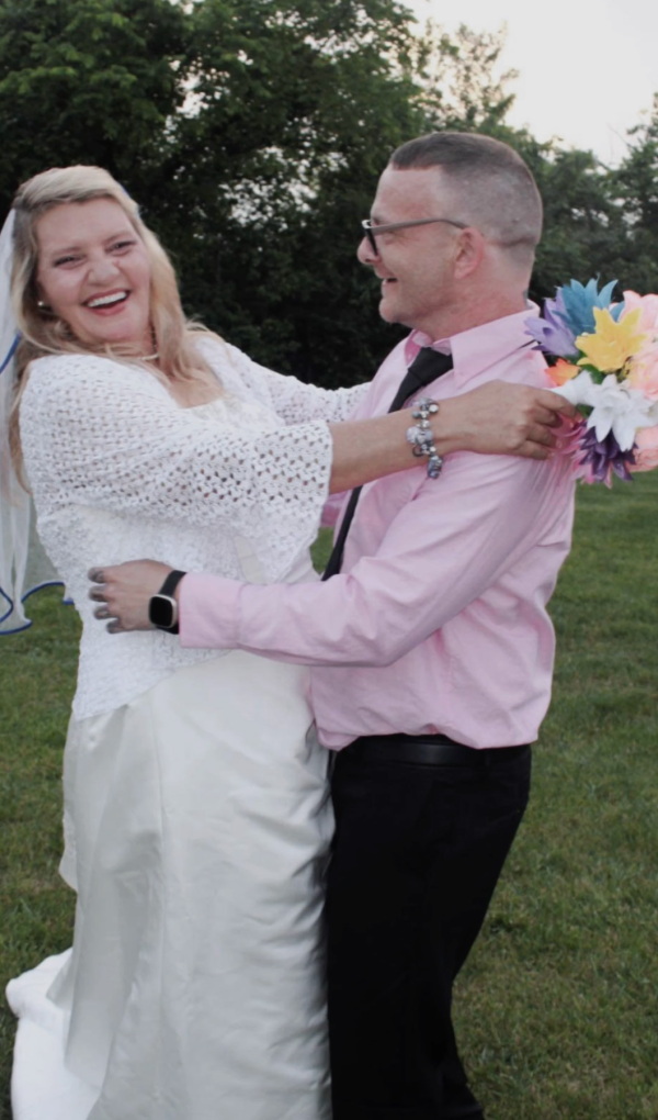 TJ Hoover danced with his sister, Donna, on her wedding day in May 2023. Donna has long blond hair and is wearing a white wedding dress. TJ is wearing a pink dress shirt and black pants. She has a bouquet in her hands. They are outside, dancing on green grass near trees.