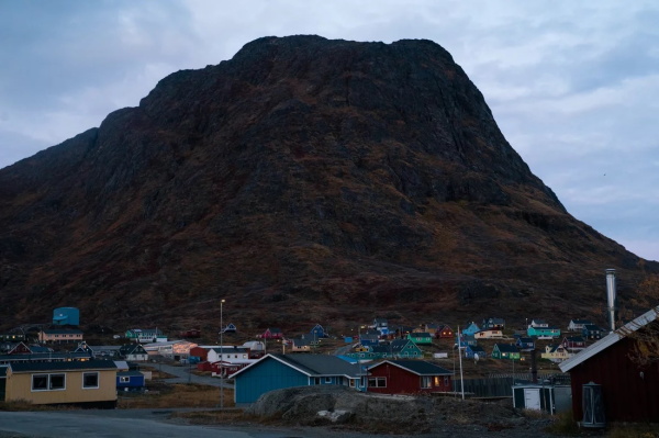 This photo shows the town of Narsaq in Greenland. Various colorful houses and buildings are in the foreground, and a large rocky mountain rises in the background.