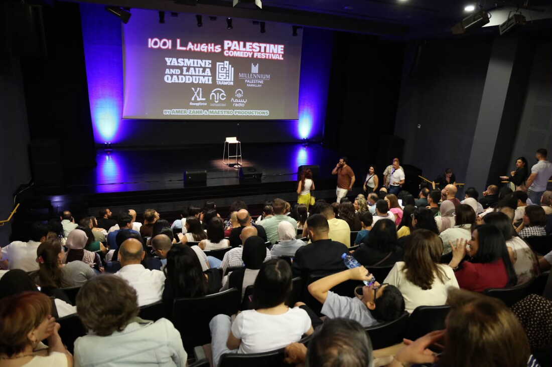 Palestinian audience members sit in rows of theater seats in an auditorium in Bethlehem to see comedians from the U.S. and other countries perform live on Zoom. A stage faces the seats, and a large screen hangs at the back of the stage. The screen says, among other things, "1001 Laughs Palestine Comedy Festival."