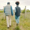Photographed from the rear, a man and a woman walk in a grassy area on a farm, with slopes rising up in the background.