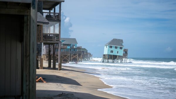 A teal house on stilts being pulled into the water. Other homes are behind it still on the sand as the water crashes against the shore.
