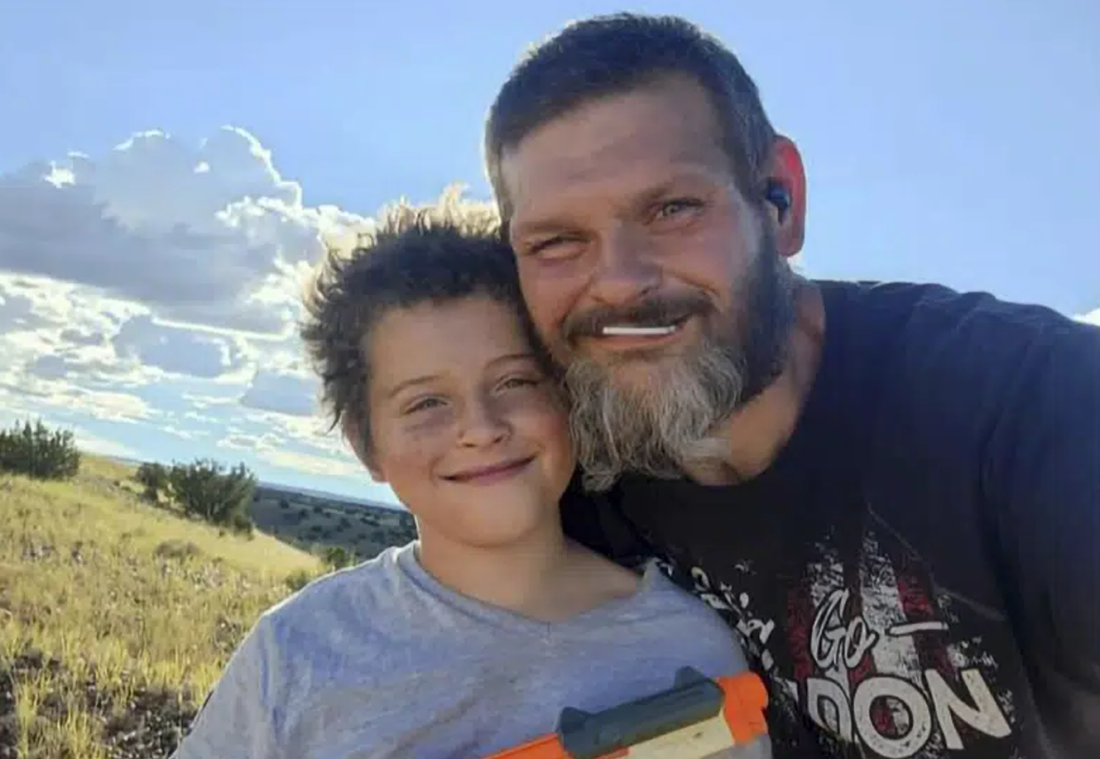 Jakob Blodgett, 9, (left) smiles in a selfie with his father, Richard Blodgett, who is hugging Jakob. Jakob is wearing a gray t-shirt and Richard is wearing a t-shirt, has a salt and pepper beard and short dark hair. There is a meadow behind them.