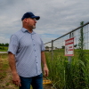 A man wearing a baseball cap stares into a lot with tall grass and a fence that says "no trespassing." 