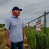 A man wearing a baseball cap stares into a lot with tall grass and a fence that says "no trespassing."