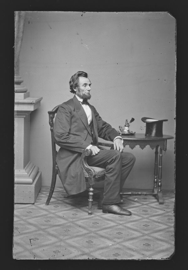 This glass plate collodion negative, a type of photograph, is of President-elect Abraham Lincoln, taken on Feb. 24, 1861, by Alexander Gardner in the days following Lincoln’s arrival in Washington, D.C., ahead of his inauguration. He is dressed in the outfit that would become his most recognizable look: a black frock coat, matching waistcoat and trousers, a narrow black cravat, and polished black leather shoes. His hat rests on the table next to him.