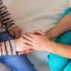 A doctor wearing scrubs and a stethoscope holds the hand of a child to provide reassurance during a medical visit.