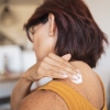 A woman applies cream to the back of her shoulder.