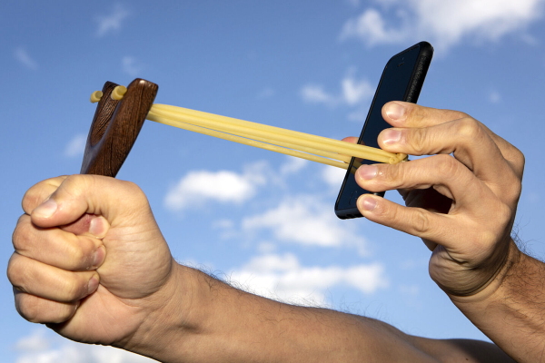A photograph shows a close-up of a hand in front of a backdrop of blue sky and white clouds holding a slingshot that is about to shoot a smart phone off into the distance.