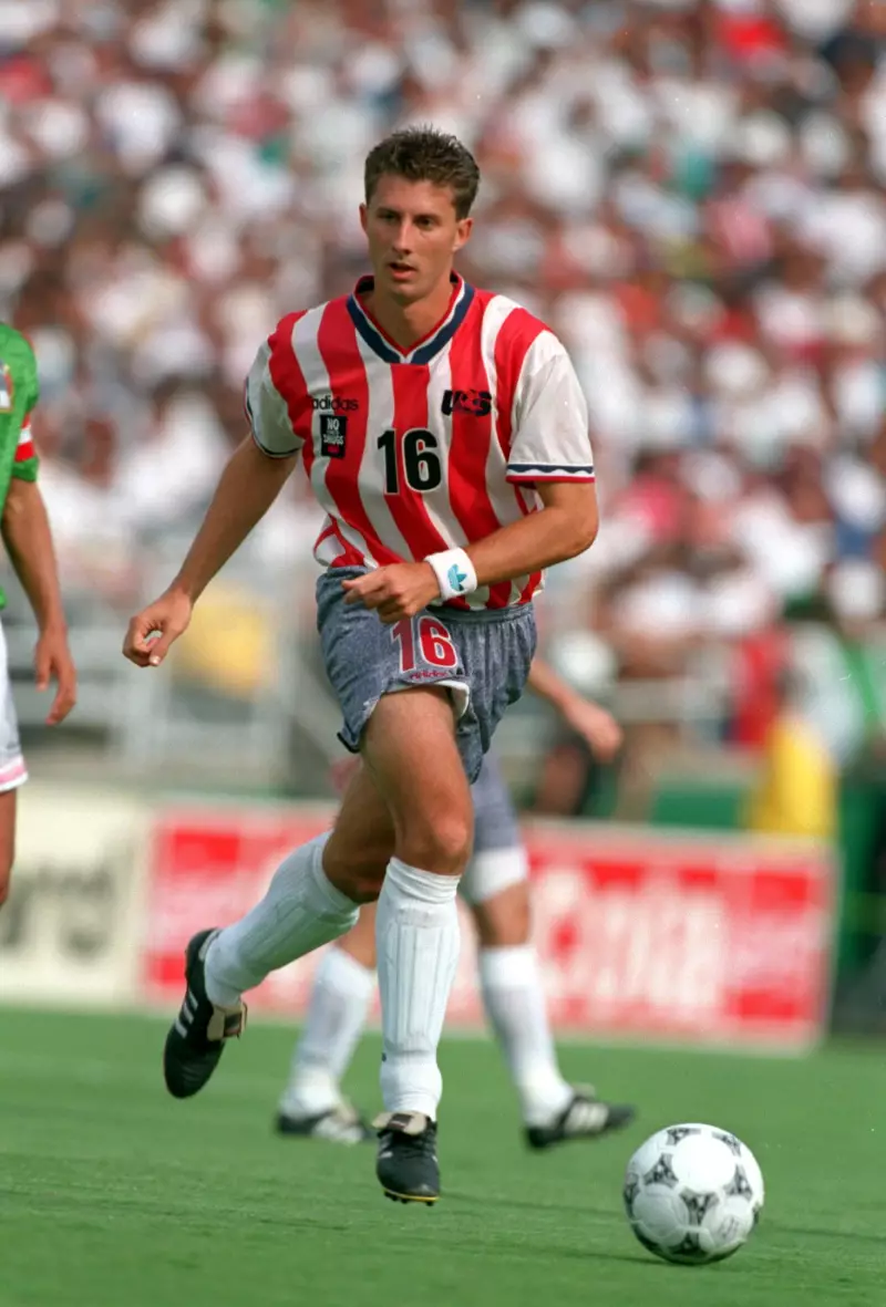 Wearing a soccer uniform, Mike Sorber plays soccer during an exhibition game at the Rose Bowl stadium in 1994. A soccer ball is on the green soccer field in front of him, and a couple of other players are in the vicinity.