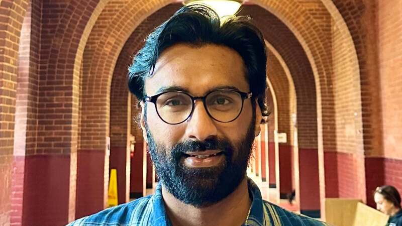 Badar Khan Suri is earing glasses and a blue plaid shirt and standing in a hallway of arches.