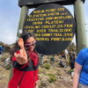 A vibrant young woman is shown in front of a sign for Kilimanjaro National Park, Tanzania. She is looking over her sunglasses and gesturing at the camera with a finger puppet on her index finger.