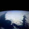 This photo of Greenland, taken from very high above, shows the outline of Greenland, which is covered in white ice and surrounded by blue ocean.