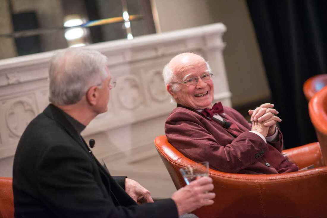 Martin Marty sits smiling in a chair, wearing a burgundy suit and bowtie, while in conversation with someone else.