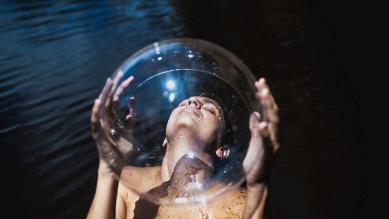 Luiza Brina nude in a river, holding a giant glass ball in front of her obscure body. 
