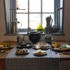 A table setting of a meal typical for working-class people in 18th and 19th-century Annapolis is displayed at Hogshead Trades Museum. A wooden table has two place settings along with plates of flat breads, small pies, large ceramic mugs and a cast iron pot situated underneath a window. Metal carafes and a green glass vase sit on the window sill.