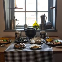 A table setting of a meal typical for working-class people in 18th and 19th-century Annapolis is displayed at Hogshead Trades Museum. A wooden table has two place settings along with plates of flat breads, small pies, large ceramic mugs and a cast iron pot situated underneath a window. Metal carafes and a green glass vase sit on the window sill.