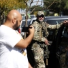 A man hold his phone up next to a Border Patrol agent dressed in fatigues and resting one arm on a vehicle parked at the curb.