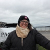 A young woman with dark hair and glasses bundled up against the cold is shown standing at her farmer's market stand. Open water and a cloudy sky are visible in the background. 
