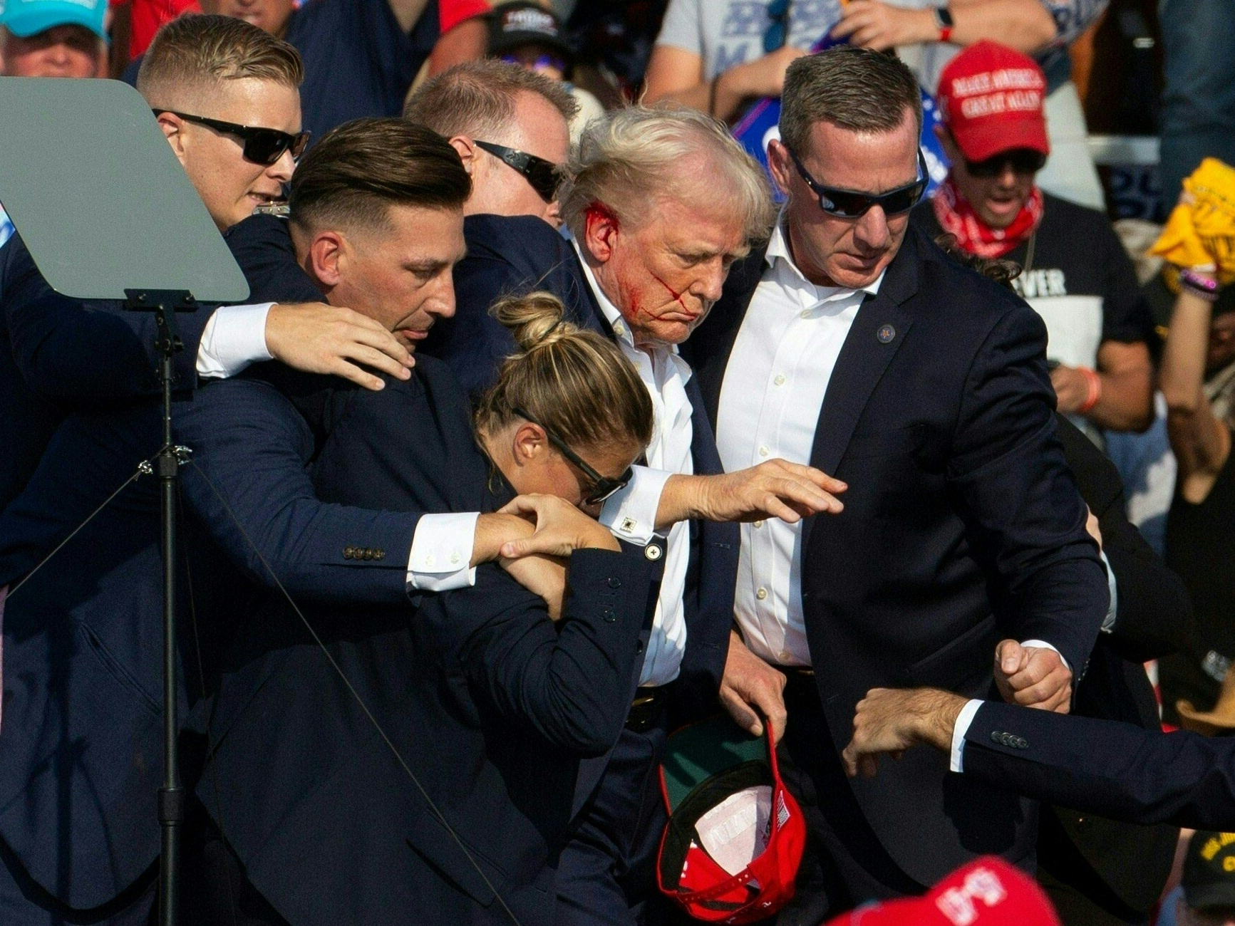 Donald Trump has blood on his face and right ear as Secret Service agents in dark suits surround him and help him off the stage amid the assassination attempt.