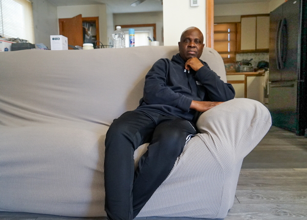 Nursing assistant Kwesi Ablordeppey sits on a grey couch wearing dark clothing.