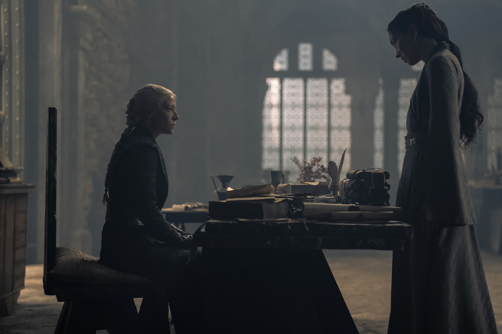 Two women (Rhaenyra, left and Mysaria, right) face each other in a large room. Rhaenyra sits at a desk covered in books, cups, and dried flowers. Mysaria stands in front of the table, and it is really dark.
