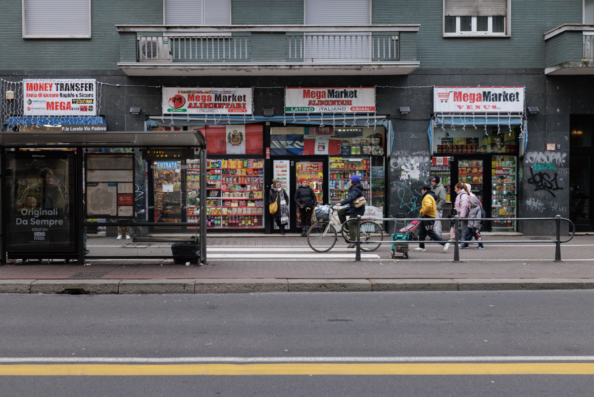 On a gray afternoon, people pass ethnic grocery shops and a money transfer center on Via Padova near Piazzale Loreto — a hub for Milan's migrant communities.